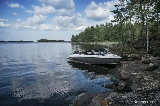 A white boat moored on rocky shores beside calm water, surrounded by trees and a bright sky with fluffy clouds.